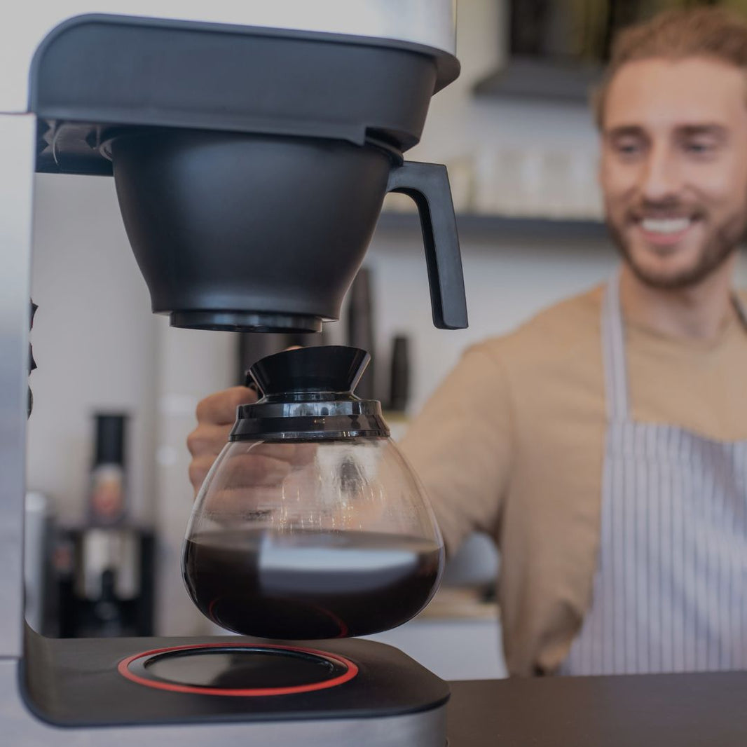 Person pouring coffee from a glass carafe into a black coffee maker with a blurred background
